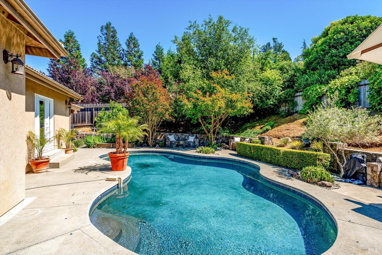 A curved pool beside a single-story home, framed by mature trees, clipped hedges and stone garden walls, in Napa's Browns Valley