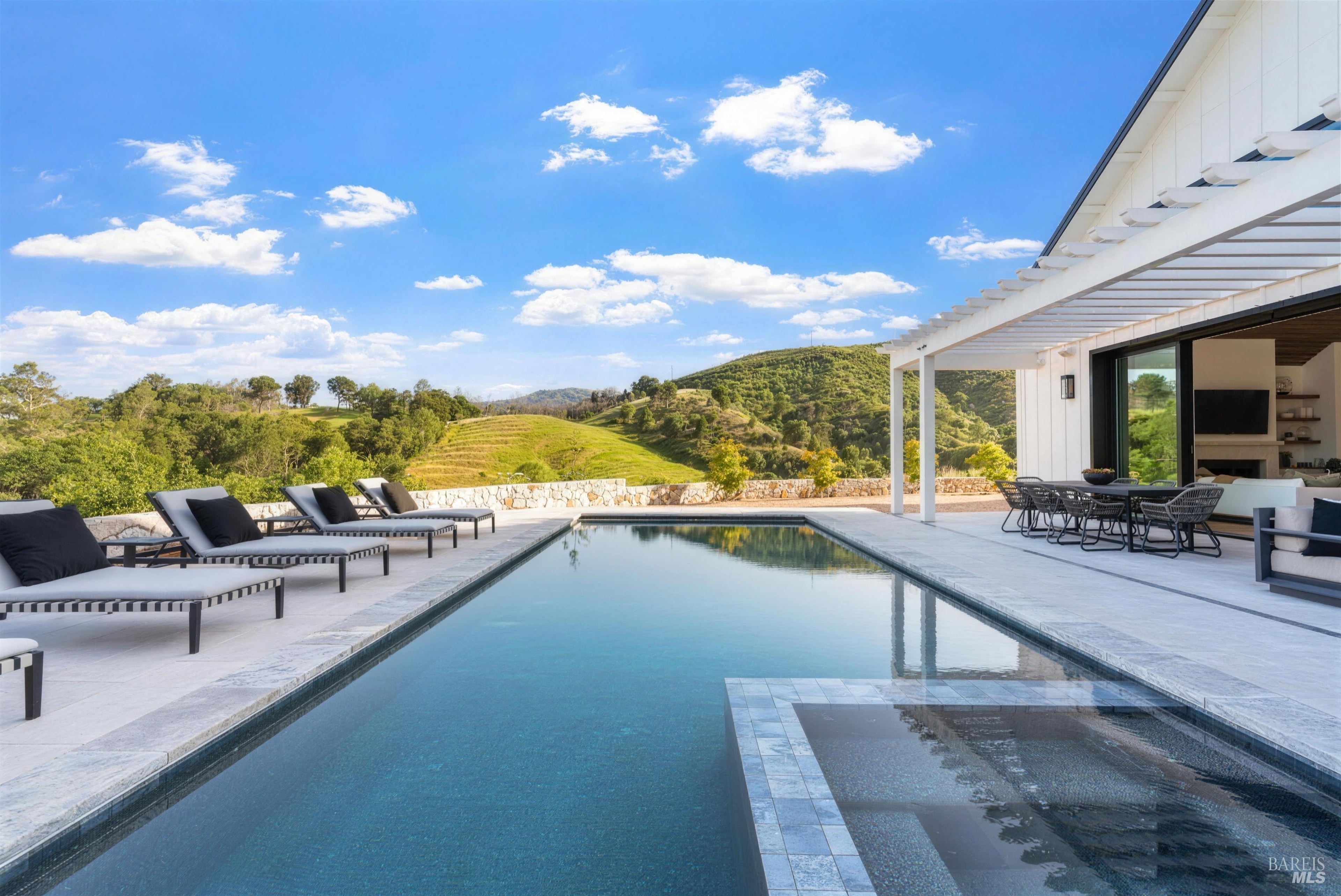 A long lap pool on a limestone deck beside a white modern farmhouse, looking out to green St. Helena hills under a blue sky